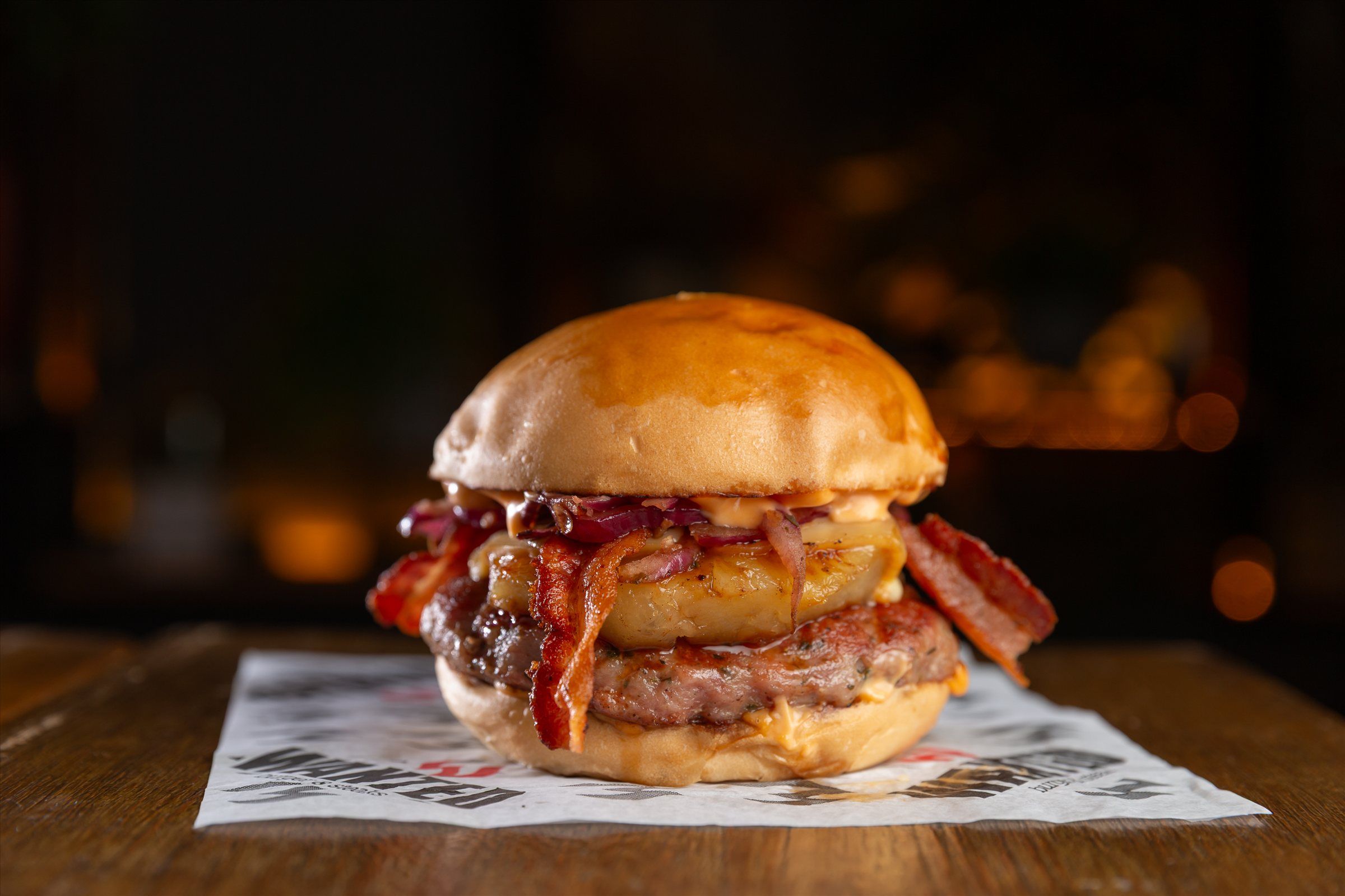 Burger com bacon e cebola caramelizada sobre o papel da Wanted, luzes âmbar do salão ao fundo